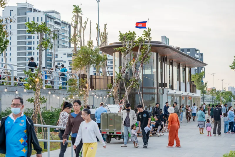 People strolling Norea City's completed riverside promenade at dusk past a pavilion with festoon lighting and the Cambodian flag